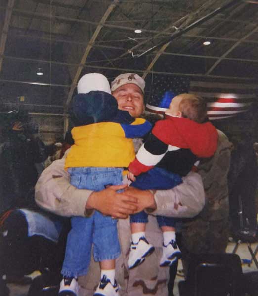 A United States Army soldier in uniform hugging two young children inside a hangar with a large American flag behind them - Tom Smoot hugs his kids during a homecoming ceremony