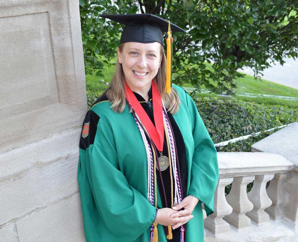 A photo of Angela Peacock in full Masters cap and gown regalia standing at the steps of University in St. Louis in May 2019. Photo courtesy of Sarah Townsend