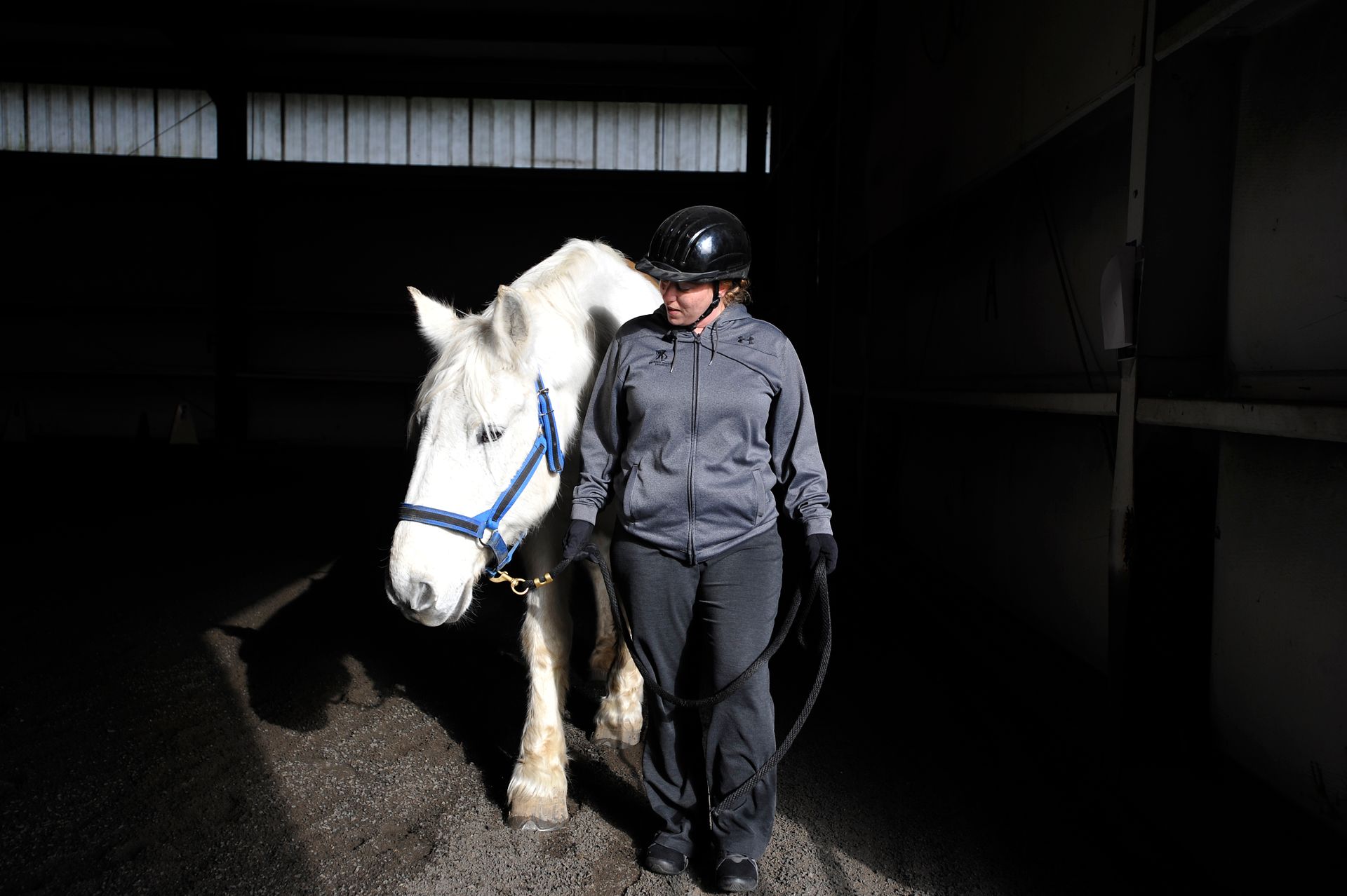 Angela, in a black riding helmet and grey Wounded Warrior Project hoodie, leads a white horse in a b