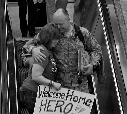 Black and white photo of woman holding a large, "Welcome Home" sign embracing a uniformed service member riding a down escalator in an airport, photo by Gina Davison