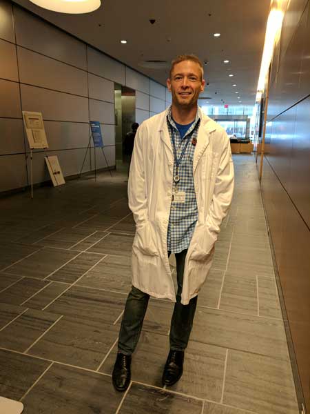 The author wears his lab coat for the first time after completing his engineering degree in September 2017. Photo courtesy of the author. A photo of the author standing in his white lab coat in a hallway, after completing his engineering degree in September 2017.