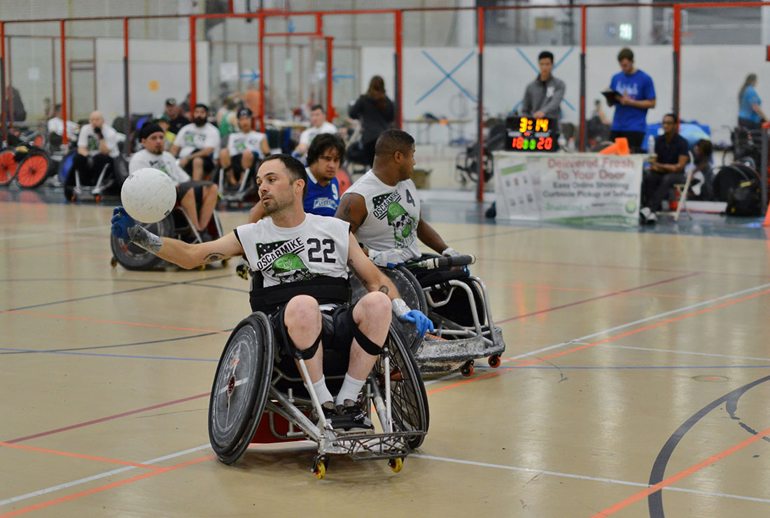 Three men in wheelchairs and white OscarMike jerseys on a basketball court play quad rugby