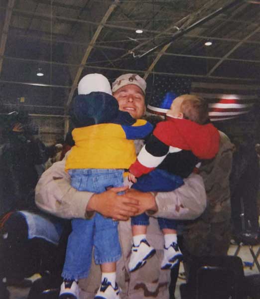 Tom Smoot hugs his kids during a homecoming ceremony in February 2004 after serving in Iraq. Photo courtesy of the author. A United States Army soldier in uniform hugging two young children inside a hangar with a large American flag behind them - Tom Smoot hugs his kids during a homecoming ceremony