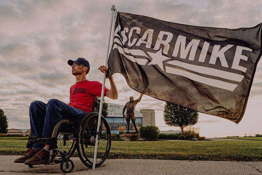 Man in a red shirt seated in a wheelchair holds a black flag with the OscarMike logo looks into the distance