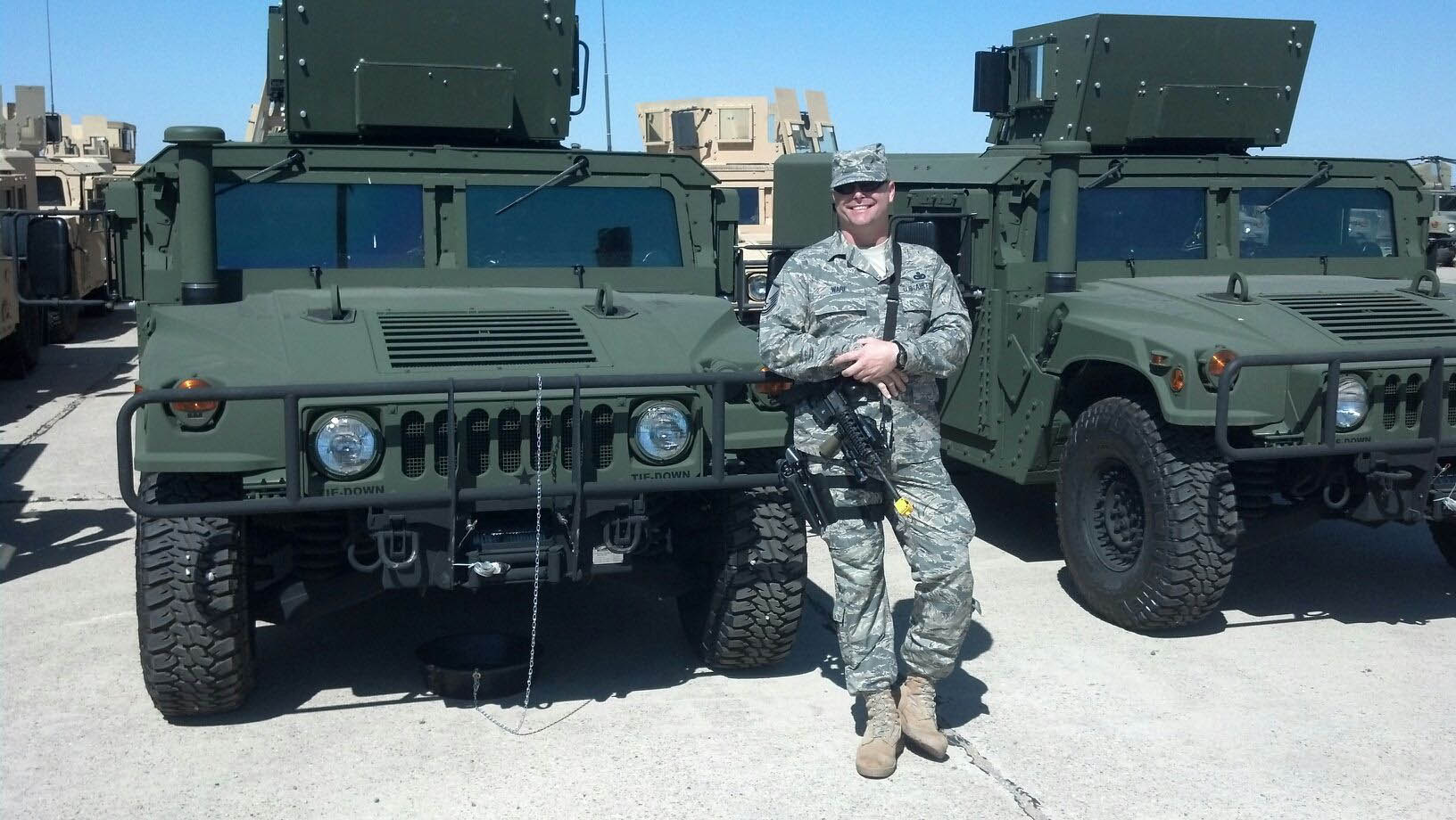 Photo of a MSgt Ware in uniform, with his weapon slung over his shoulder, front of green armored Humvees