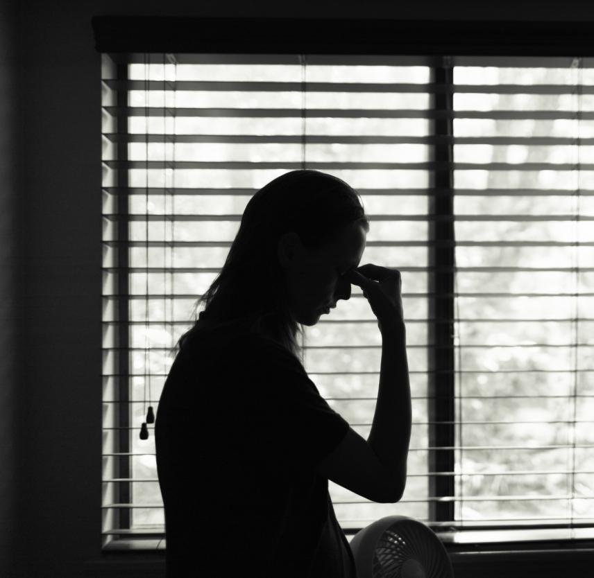 A woman stands alone in a darkened room, holding her head in her hand