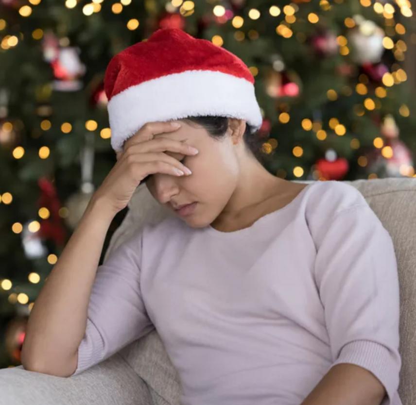 Woman in a red and white Santa hat seated, cradling her head in her hand, in front of a lighted Christmas tree