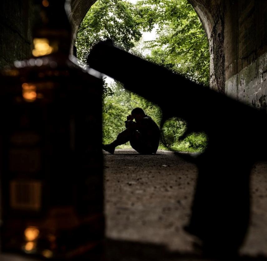 A person sits in a dark tunnel, their head in their hands, a gun and liquor bottle in the foreground