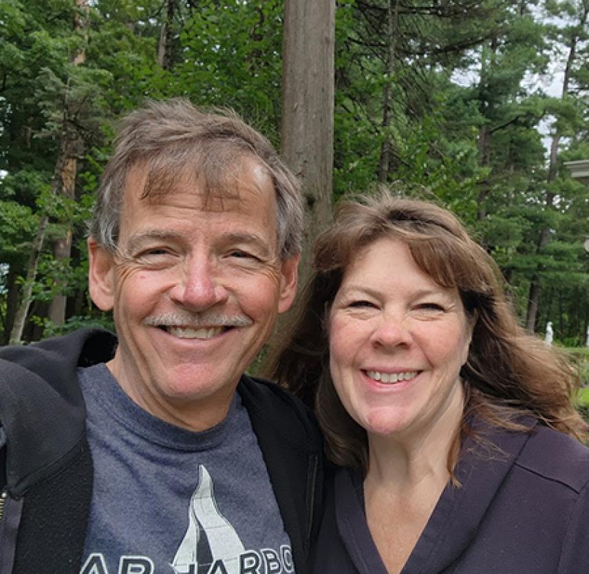 David and his wife smiling in front of tall pine trees