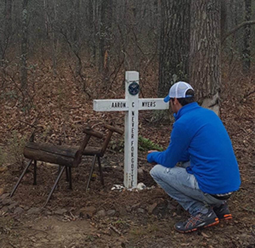 Steven crouches by Aaron's white memorial cross in the woods by two wooden deer sculptures