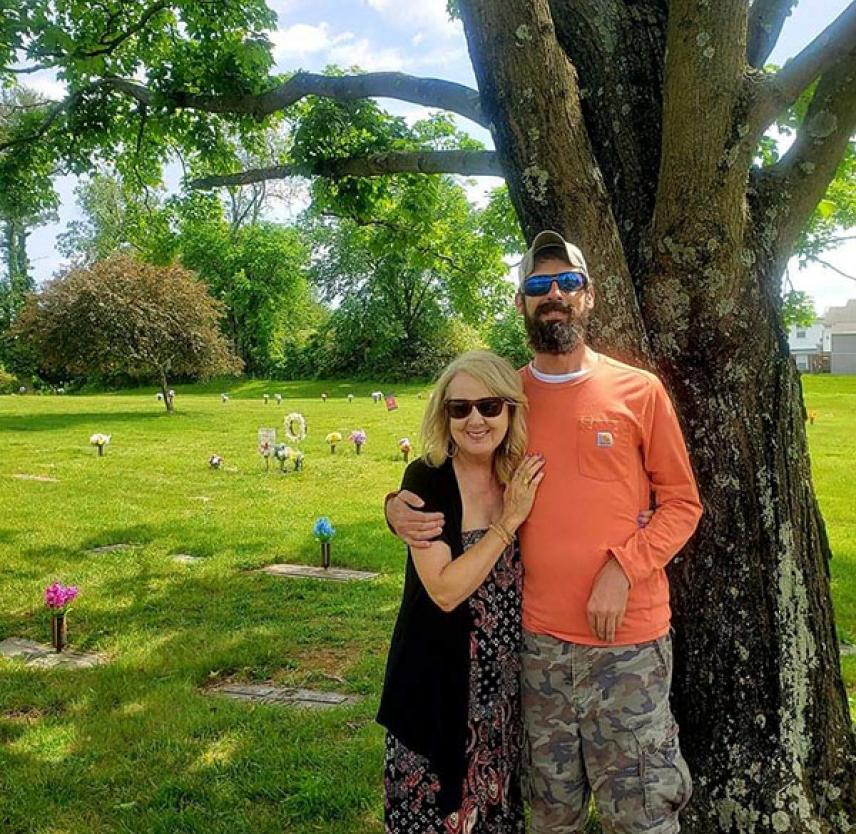 Norma and Steven smiling in front of a large tree at the cemetery