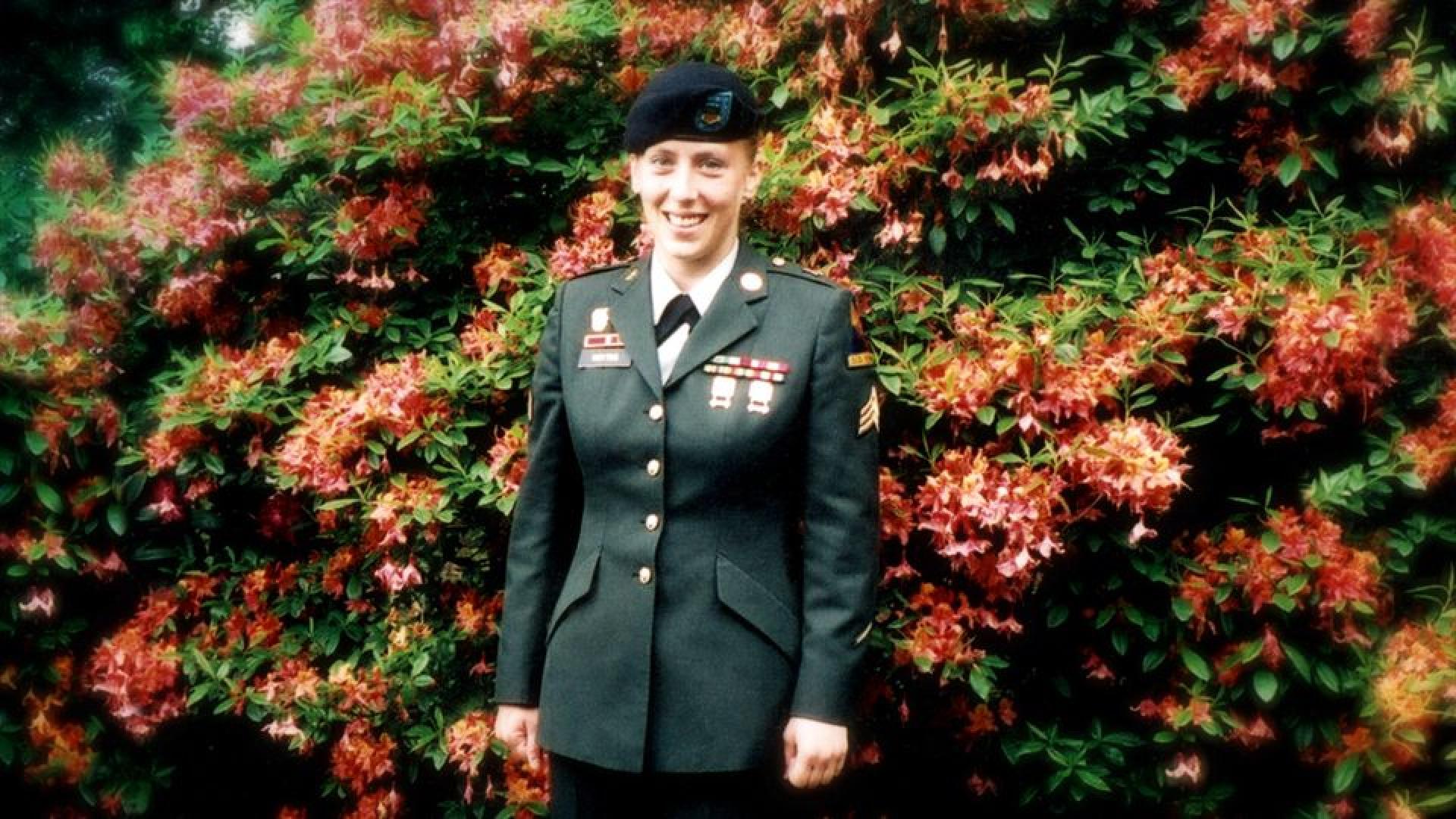 Angela Peacock, a young woman, stands in her Army dress uniform in front of a tall bright red and dark green azalea.