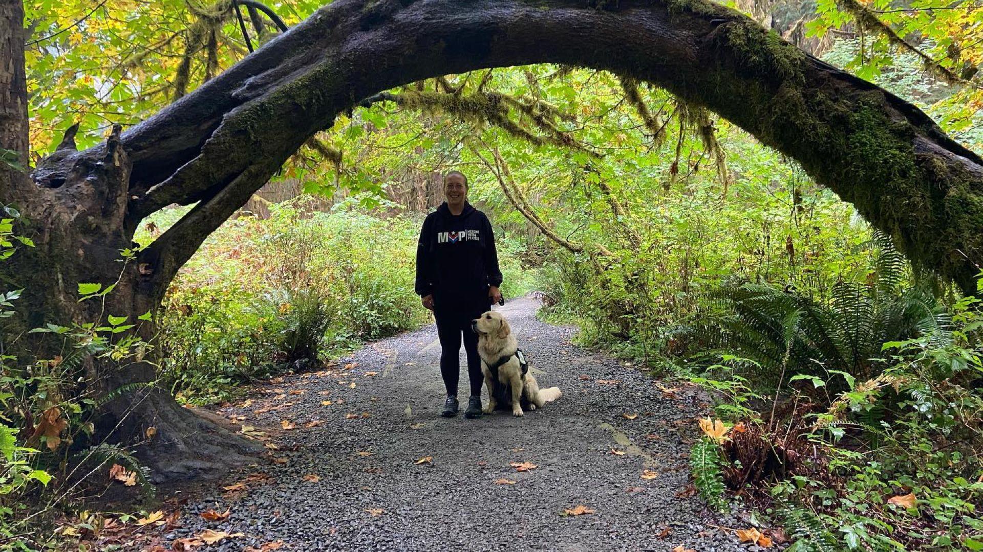 Angela and her service dog Raider stand under the arch of a fallen tree in the woods