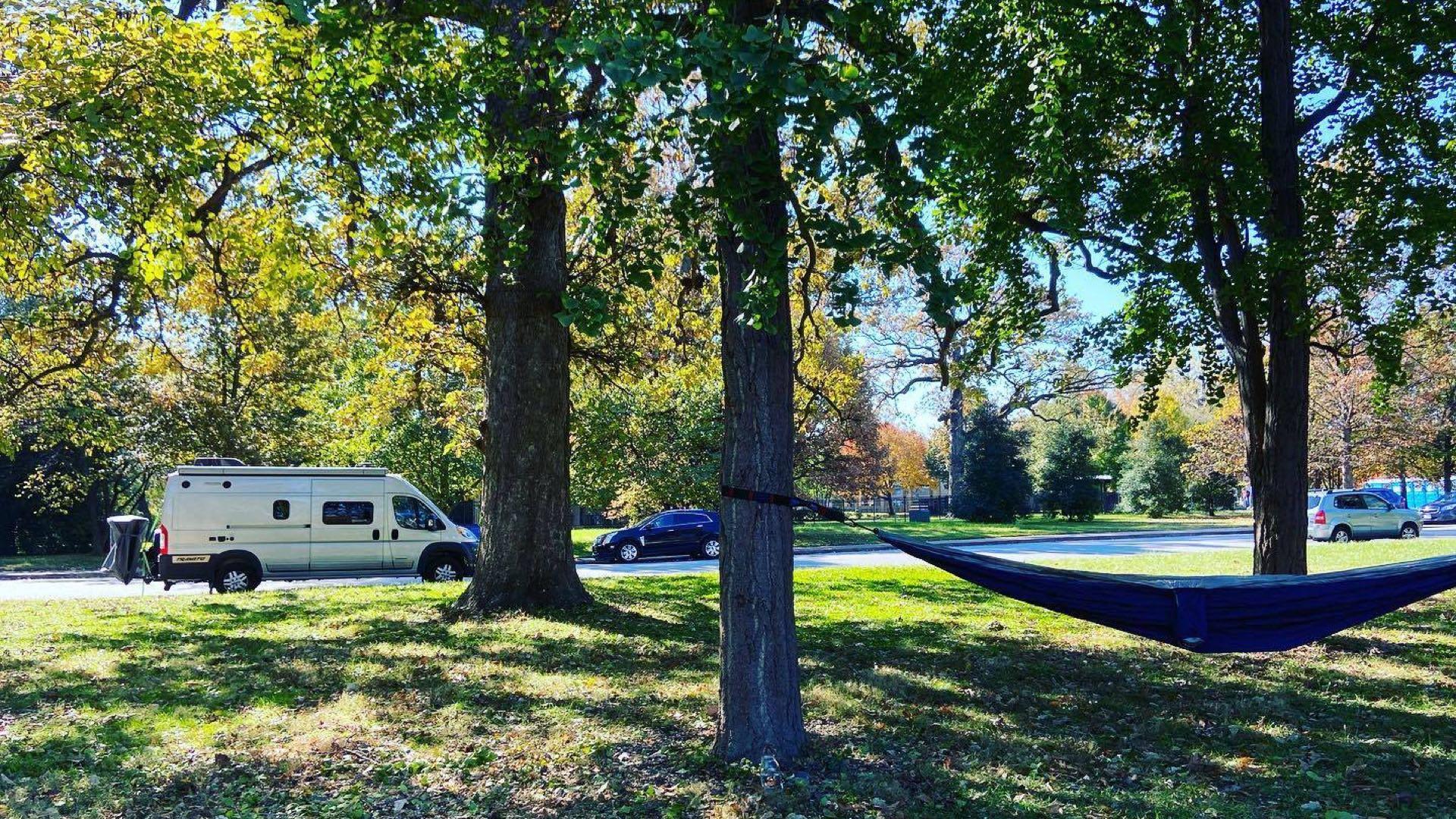 alt="Angela’s RV, left, parked by tall trees, two of which support a hammock, right"