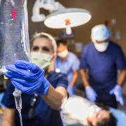 Nurse checks an IV bag while other doctors and nurses attend a patient in a coma