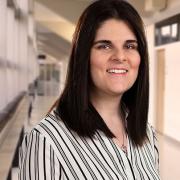 Ashton Kroner smiles in a hallway with shoulder-length brown hair, wearing a striped shirt