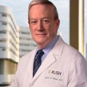 Professional headshot of Dr. Robert Shulman smiling at the camera, wearing a white doctor's coat, with a tall building in the background