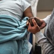 One black man holds the hand of an older black man in a hospital bed