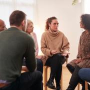 A small support group sit in a circle in a white room, talking and listening intensely
