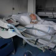 Patient lies in bed with a tracheostomy showing air tubes and machinery