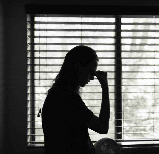 A woman stands alone in a darkened room, holding her head in her hand