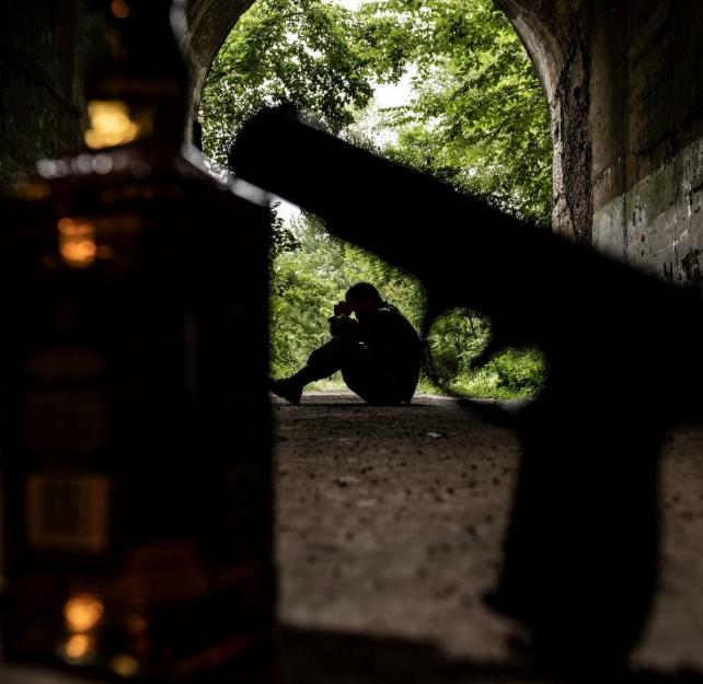 A person sits in a dark tunnel, their head in their hands, a gun and liquor bottle in the foreground