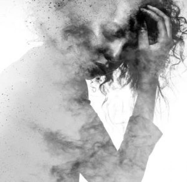 Greyscale image - A woman in a suit cradles her head, with a pained look on her face, obscured by pa