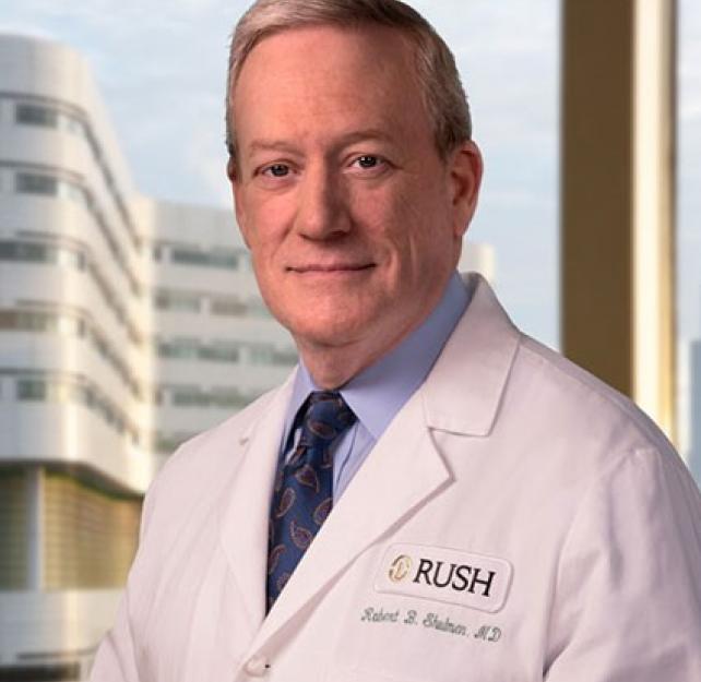 Professional headshot of Dr. Robert Shulman smiling at the camera, wearing a white doctor's coat, with a tall building in the background