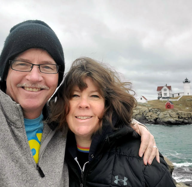 David Grant and his wife smiling at the camera in front of a lighthouse