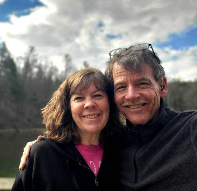 David and his wife smiling in front of pine trees on a sunny day