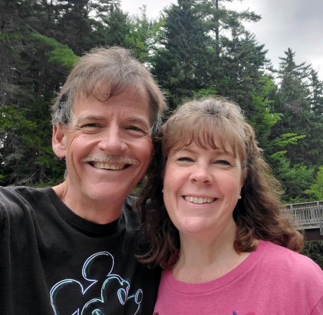 David Grant and his wife smiling in front of tall pine trees