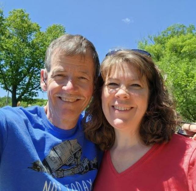 David wearing a blue t-shirt and his wife in a red shirt smiling outside in the sun