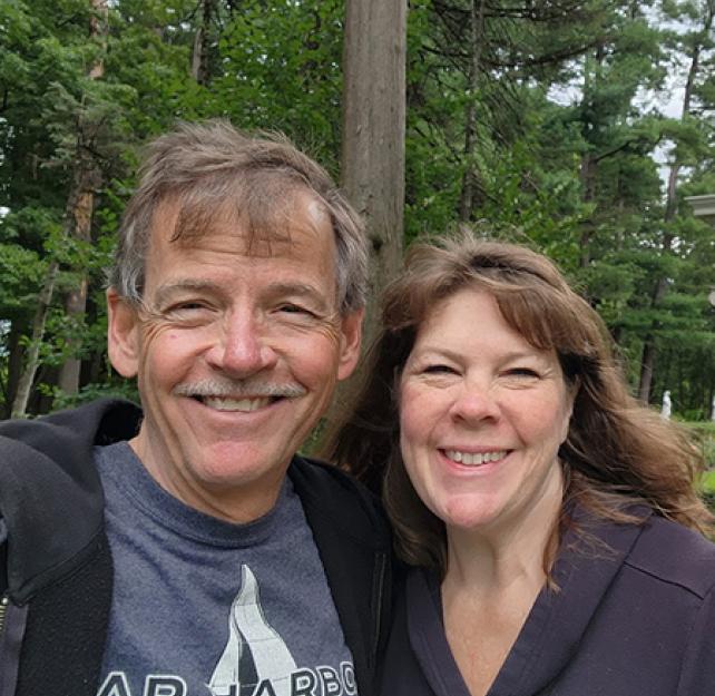 David and his wife smiling in front of tall pine trees