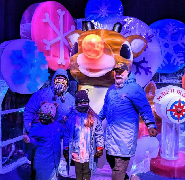 Stacey, Russ, and their daughters smiling in front of a giant ice sculpture of Rudolph the Red Nose 