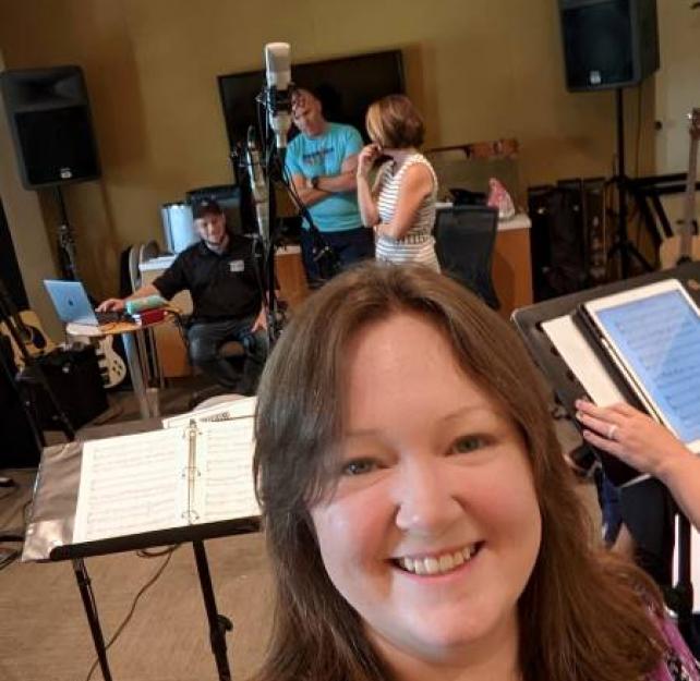 Stacey Shade-Ware smiles at the camera with music stands, a microphone, and others in the background