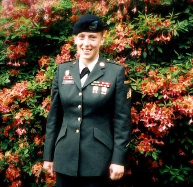 Angela Peacock stands in her Army dress uniform in front of a tall bright red and dark green azalea.