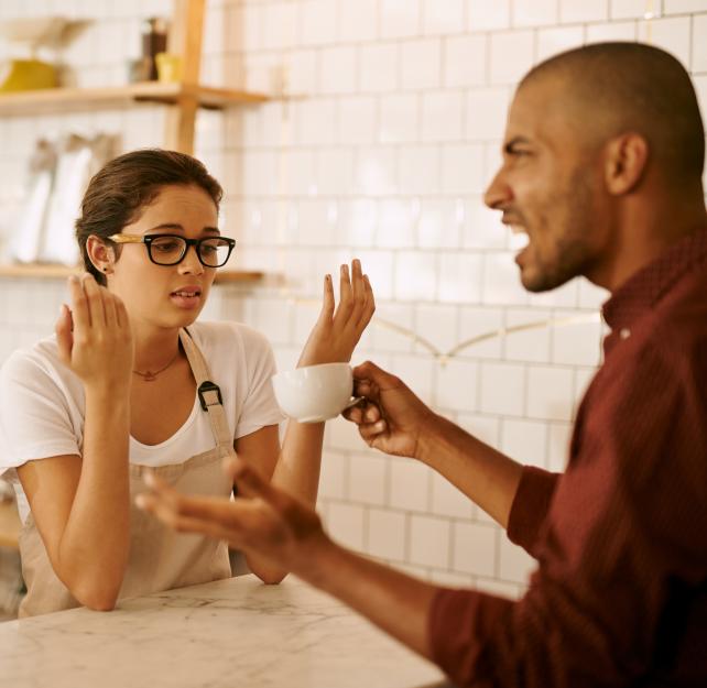 Photo of a man holding a coffee cup yelling at a woman wearing glasses and a tan apron
