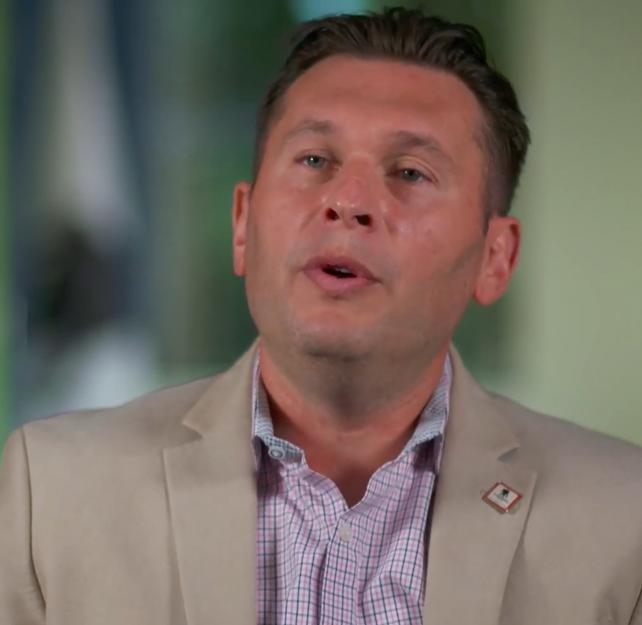 Screenshot of Dr. Alexander Balbir in a tan suit with a green-blue background