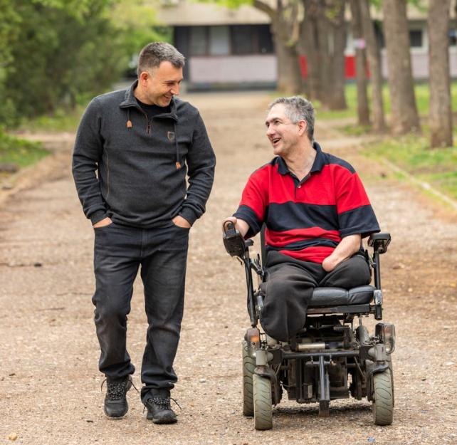 Photo of two men, one walking, one driving his motorized wheelchair down a tree-lined gravel path towards the camera, with a house in the background