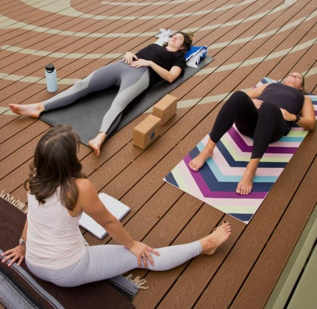 Two women lie on yoga mats while one seated instructor leads a meditation