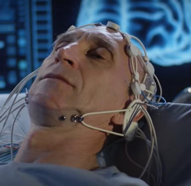 Screenshot of a male patient getting a brain scan with a digital image of a brain behind him