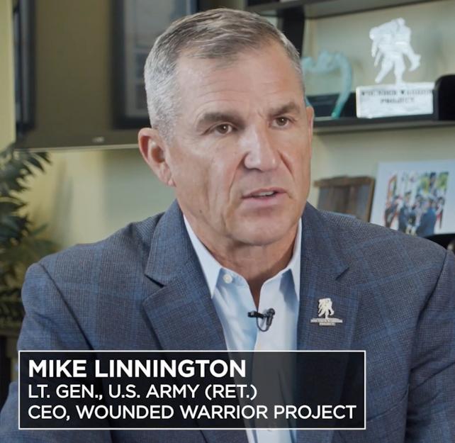 WWP CEO Mike Linnington wearing a blue suit sits in his office with Wounded Warrior Project memorabilia on the shelves behind him