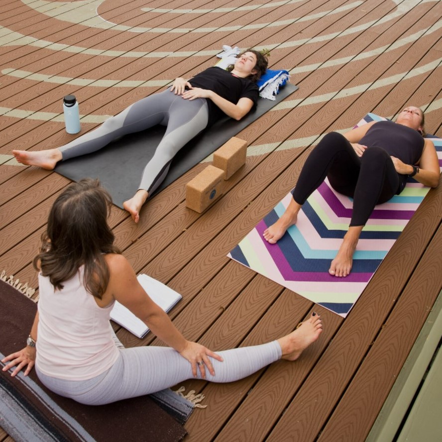 Two women lie on yoga mats while one seated instructor leads a meditation