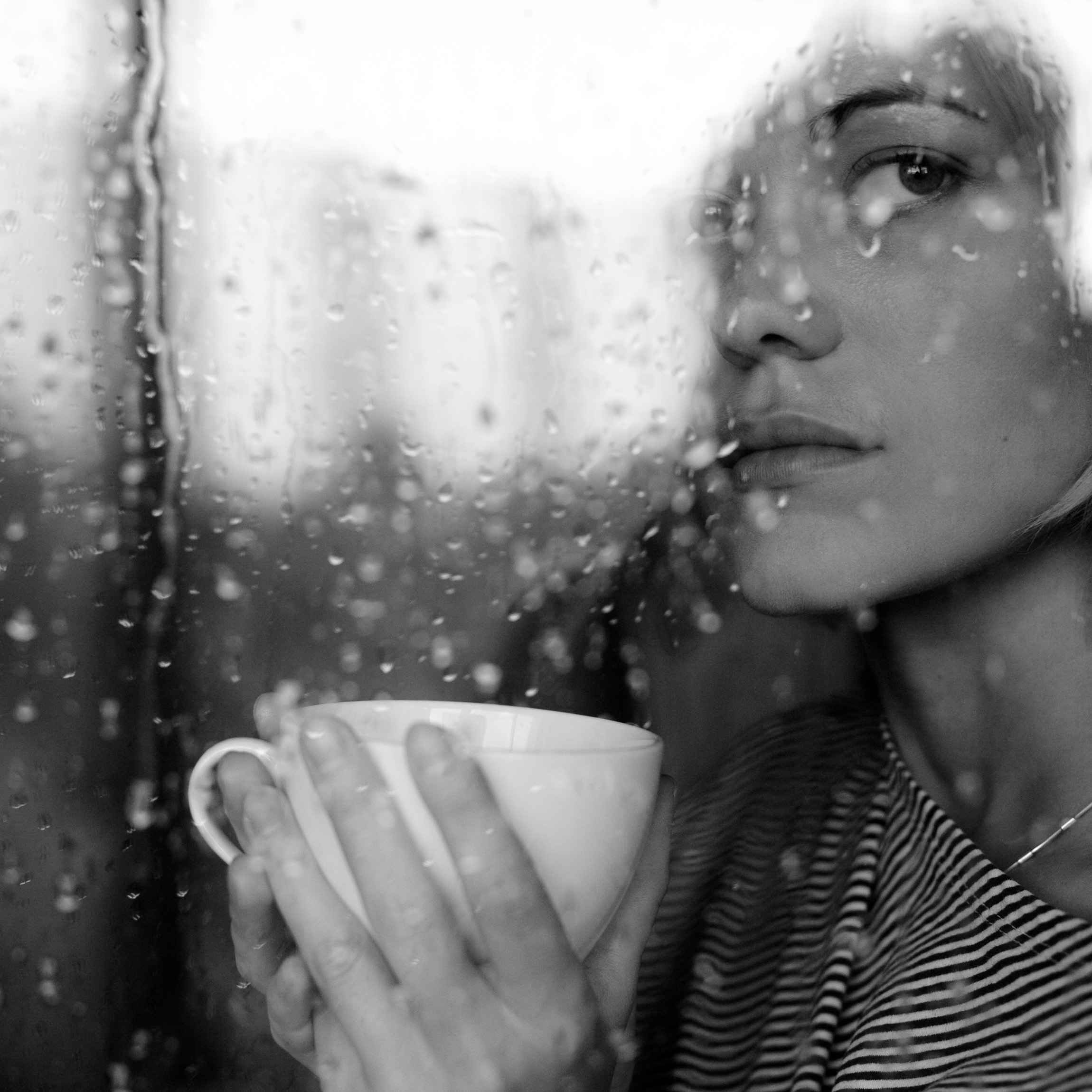 Woman looking out a rainy window while holding mug.